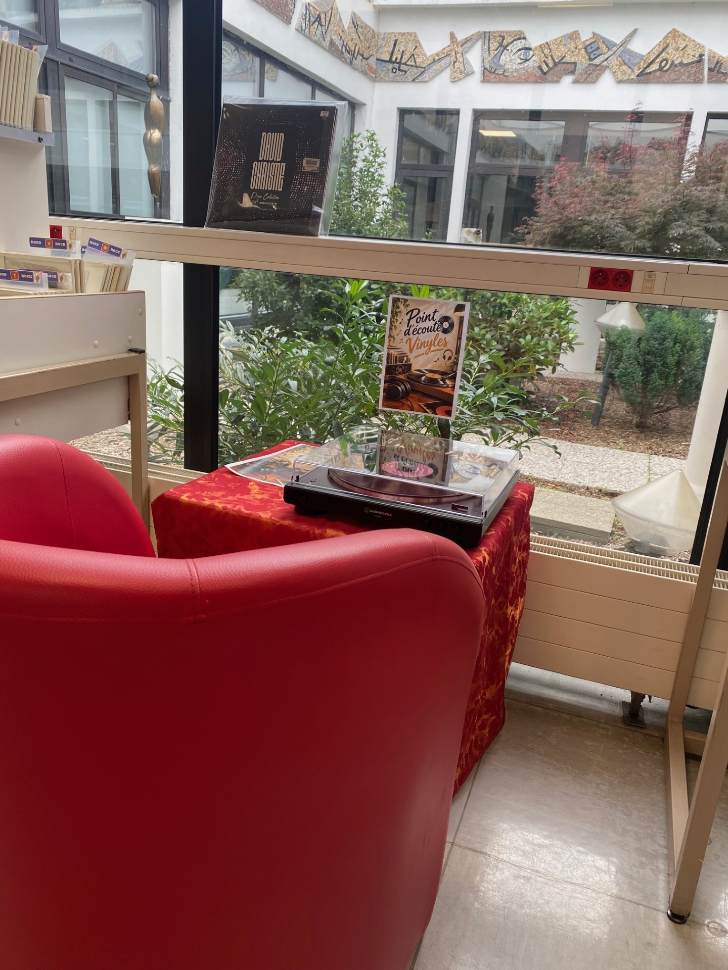 Un fauteuil rouge au premier plan, une petite table avec un tissu rouge sur lequel est posée une platine vinyle. Au second plan, nous voyons un jardin d'agrément.